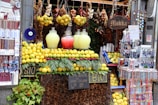 A vibrant market stall featuring an array of lemons and jugs of lemonade, flanked by hanging spices, garlic, and decorative items. Colorful beads and ornaments are displayed on the side, with signs indicating organic products for sale.