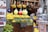 A vibrant market stall featuring an array of lemons and jugs of lemonade, flanked by hanging spices, garlic, and decorative items. Colorful beads and ornaments are displayed on the side, with signs indicating organic products for sale.