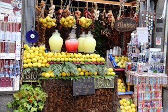 A vibrant market stall featuring an array of lemons and jugs of lemonade, flanked by hanging spices, garlic, and decorative items. Colorful beads and ornaments are displayed on the side, with signs indicating organic products for sale.