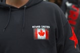 A black hoodie features a patch with the Canadian flag and the words 'Stand United' on the chest. The hoodie includes drawstrings and a red star design on the sleeve.