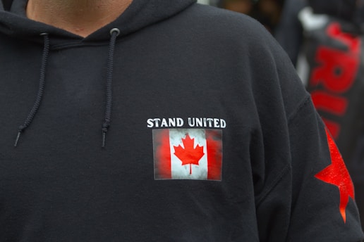 A black hoodie features a patch with the Canadian flag and the words 'Stand United' on the chest. The hoodie includes drawstrings and a red star design on the sleeve.