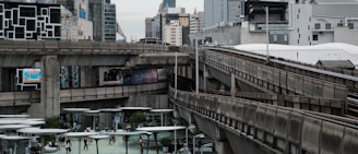 An urban scene showcasing a cityscape with a multi-layered elevated train system. The foreground features a train on the track, surrounded by modern buildings. Below, a public space with people walking and modern architectural structures is visible. The background consists of tall, varied buildings, conveying a bustling metropolitan environment.