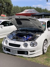 A white car is parked with its hood open, revealing the engine. The car is positioned near a grassy area with trees in the background. A red and white tent with the word 'Toyota' is visible nearby. Another white car is partially visible next to the main vehicle.