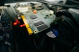 A close-up image of a computer's internal components, including a circuit board with several black chips and a gold connector. The background includes additional hardware components, wires, and cables with a fan partially visible.