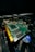 A close-up view of a computer graphics card with visible electronic components and connectors. The circuit board is green with gold connectors and several black chips. Part of a cooling fan with the Intel logo is visible beneath the card, along with visible wiring.