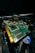A close-up view of a computer graphics card with visible electronic components and connectors. The circuit board is green with gold connectors and several black chips. Part of a cooling fan with the Intel logo is visible beneath the card, along with visible wiring.