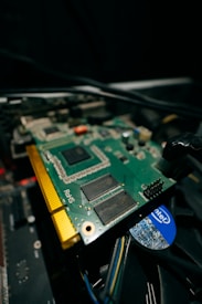 A close-up view of a computer graphics card with visible electronic components and connectors. The circuit board is green with gold connectors and several black chips. Part of a cooling fan with the Intel logo is visible beneath the card, along with visible wiring.