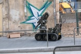 A robotic vehicle with caterpillar tracks is positioned on a paved sidewalk next to a graffiti-painted wall. The graffiti features a large green and blue star-like shape. There is a cardboard box lying on the ground nearby. The scene appears to be a construction or demolition site, with a yellow excavator visible in the background behind a wire fence.
