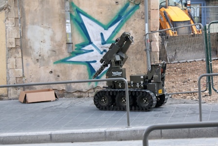 A robotic vehicle with caterpillar tracks is positioned on a paved sidewalk next to a graffiti-painted wall. The graffiti features a large green and blue star-like shape. There is a cardboard box lying on the ground nearby. The scene appears to be a construction or demolition site, with a yellow excavator visible in the background behind a wire fence.