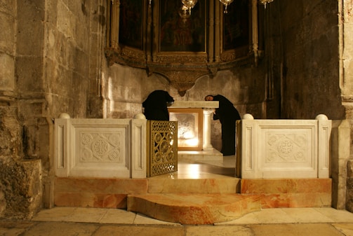 A dimly lit stone-walled room with ornate decorations and a central altar, where two people dressed in black robes are standing. The altar is made of white marble with intricate carvings and golden accents. The atmosphere is somber and historic.