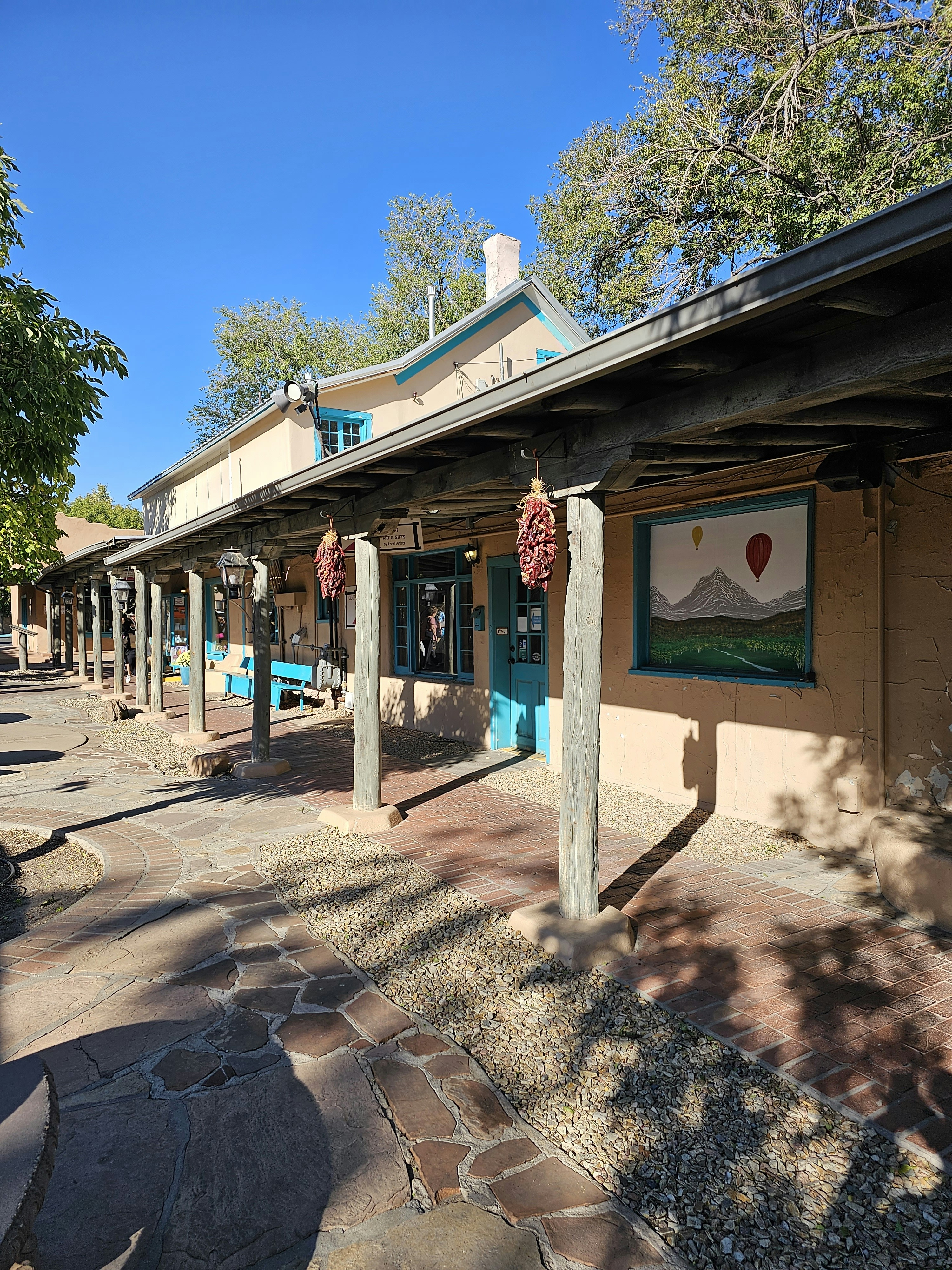 A rustic building with adobe-style architecture featuring wooden beams and columns. The building facade includes windows and a door with turquoise trim. Red chili ristras hang from the beams. A large mural of mountains and hot air balloons is displayed on the wall. The ground is paved with a combination of brick and stone, and there are trees in the background with a clear blue sky.