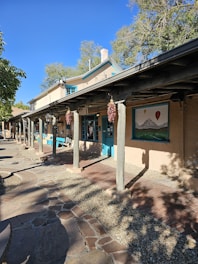 A rustic building with adobe-style architecture featuring wooden beams and columns. The building facade includes windows and a door with turquoise trim. Red chili ristras hang from the beams. A large mural of mountains and hot air balloons is displayed on the wall. The ground is paved with a combination of brick and stone, and there are trees in the background with a clear blue sky.