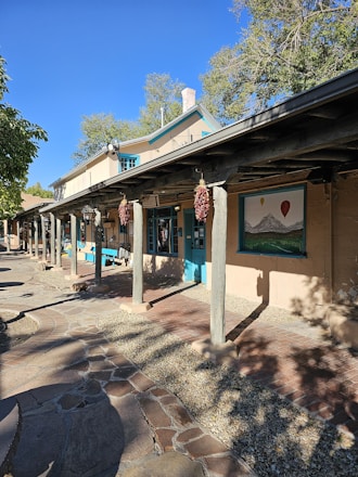 A rustic building with adobe-style architecture featuring wooden beams and columns. The building facade includes windows and a door with turquoise trim. Red chili ristras hang from the beams. A large mural of mountains and hot air balloons is displayed on the wall. The ground is paved with a combination of brick and stone, and there are trees in the background with a clear blue sky.