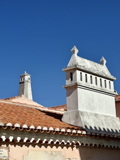 Two chimneys stand out against a clear blue sky, each uniquely designed with detailed masonry work. The rooftops are covered with traditional terracotta tiles, creating a warm contrast with the bright sky. A street sign is partially visible in the lower left corner.