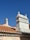 Two chimneys stand out against a clear blue sky, each uniquely designed with detailed masonry work. The rooftops are covered with traditional terracotta tiles, creating a warm contrast with the bright sky. A street sign is partially visible in the lower left corner.