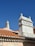 Two chimneys stand out against a clear blue sky, each uniquely designed with detailed masonry work. The rooftops are covered with traditional terracotta tiles, creating a warm contrast with the bright sky. A street sign is partially visible in the lower left corner.