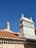 Two chimneys stand out against a clear blue sky, each uniquely designed with detailed masonry work. The rooftops are covered with traditional terracotta tiles, creating a warm contrast with the bright sky. A street sign is partially visible in the lower left corner.