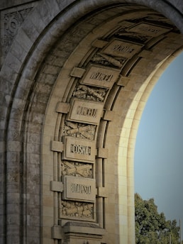 An intricately designed stone archway featuring engraved city names on rectangular plaques, surrounded by detailed ornamental architecture. The arch displays a series of relief sculptures and names including cities like Budapest. The sun casts shadows enhancing the texture of the stone, and a patch of trees is visible in the background.