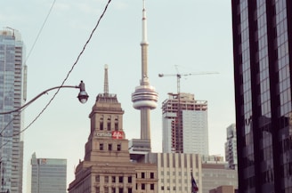 A cityscape featuring the CN Tower prominently in the center, surrounded by skyscrapers and buildings. The urban environment is complemented by construction cranes and a visible 'Canada Life' building.