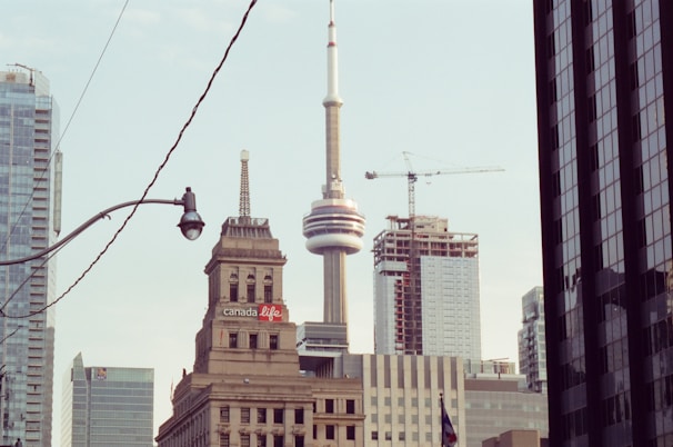 A cityscape featuring the CN Tower prominently in the center, surrounded by skyscrapers and buildings. The urban environment is complemented by construction cranes and a visible 'Canada Life' building.