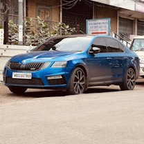 A vibrant blue sedan is parked on the side of a street. The vehicle appears sleek and modern, with black-tinted windows and distinctive alloy wheels. In the background, there is an industrial building with signage and a small amount of greenery visible.