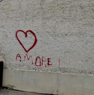 A textured wall with rough gray surface features a red heart drawn with spray paint. Below the heart, the word 'AMORE' is written in the same red paint. The lower part of the wall is smoother and shows signs of wear.