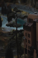 A miniature scene depicting a tall, detailed model of a tree next to a scale model of a building adorned with windows and a satellite dish. The background suggests a winding road surrounded by more model trees and landscape features.