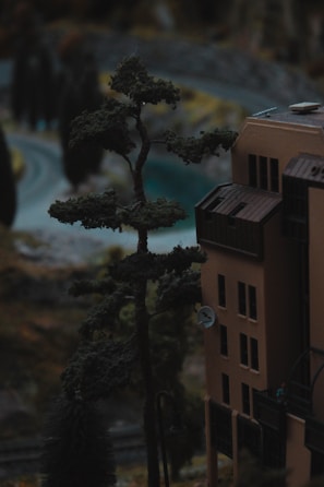 A miniature scene depicting a tall, detailed model of a tree next to a scale model of a building adorned with windows and a satellite dish. The background suggests a winding road surrounded by more model trees and landscape features.