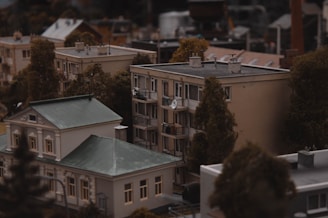 A miniature model of an urban neighborhood features detailed buildings and trees. The scene includes a multi-story residential building with balconies and a smaller structure with a green roof. Trees are strategically placed around the buildings, contributing to a realistic cityscape.