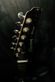A close-up view of a guitar headstock with tuning pegs. The lighting is dim, highlighting the metallic and wooden textures of the guitar.