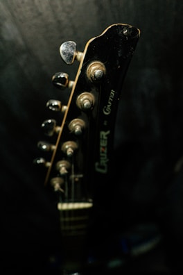 A close-up view of a guitar headstock with tuning pegs. The lighting is dim, highlighting the metallic and wooden textures of the guitar.