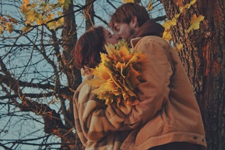 A couple sharing a tender embrace under a canopy of autumn leaves, colors warm and rich.