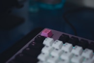 A close-up of a mechanical keyboard featuring a unique pink keycap with a symbol or logo. The rest of the keyboard comprises sleek black and white keycaps, with a subtle blue light illuminating the keys from beneath.