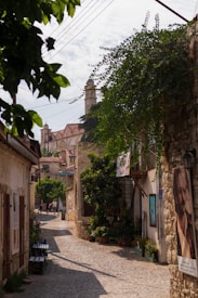 A quaint cobblestone street lined with rustic stone buildings and lush greenery. Leaves from overhanging branches create a natural frame, while colorful potted plants add vibrancy. In the background, architectural details of a church or historic building are visible, characterized by its pointed arch windows and a steeple. Posters and artwork are displayed on the walls, adding character to the narrow passageway. A couple of people walk at a distance, contributing to the serene, leisurely atmosphere.