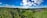 A panoramic landscape features rolling hills covered in lush greenery and dense forests under a vibrant blue sky with scattered fluffy clouds. A small village with clustered rooftops nestles on the right, surrounded by agricultural fields.