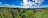 A panoramic landscape features rolling hills covered in lush greenery and dense forests under a vibrant blue sky with scattered fluffy clouds. A small village with clustered rooftops nestles on the right, surrounded by agricultural fields.