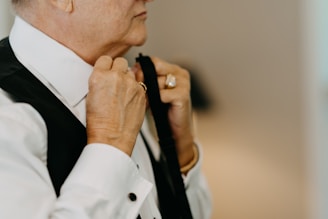 A person adjusting a dark tie around the collar of a white dress shirt. The individual is holding the tie with both hands, wearing a ring on one finger. The focus is on the hands and upper body, with a blurred background providing a soft, neutral tone.