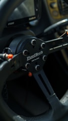 A close-up view of a black racing-style steering wheel with a visible brand logo. The design features metallic elements with a matte black finish. There are some cables and connectors visible around the central hub.