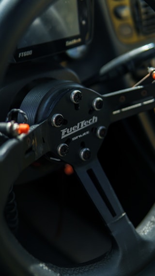 A close-up view of a black racing-style steering wheel with a visible brand logo. The design features metallic elements with a matte black finish. There are some cables and connectors visible around the central hub.