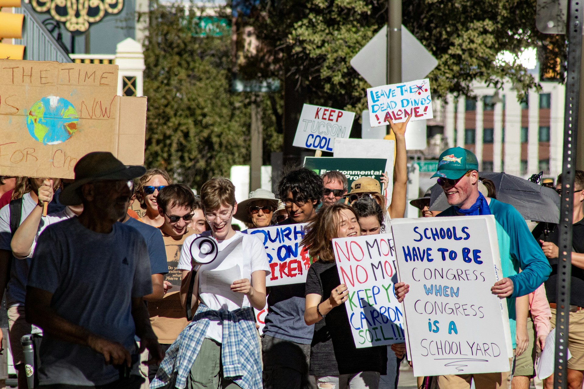 A group of people participating in a protest march, holding various signs with messages related to environmental issues. A person in the foreground is holding a megaphone, while others are smiling and actively engaging in the demonstration.