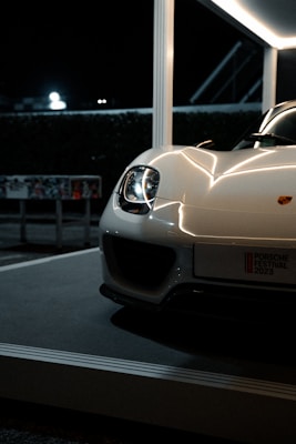 A sleek, white sportscar displayed on a platform at night, highlighting its shining headlights and smooth, glossy curves. The setting features a modern structure with bright lighting that accentuates the car's elegant design.