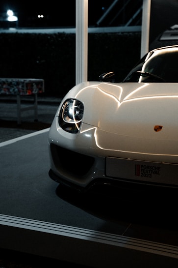 A sleek and modern sports car with a shiny white exterior and prominent headlights. The car is partially visible, positioned in a well-lit indoor setting, possibly on display or at an event. The logo is visible on the front, and a license plate with event details is mounted below.
