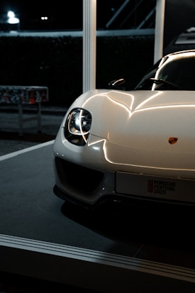 A sleek and modern sports car with a shiny white exterior and prominent headlights. The car is partially visible, positioned in a well-lit indoor setting, possibly on display or at an event. The logo is visible on the front, and a license plate with event details is mounted below.
