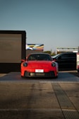 A bright red sports car is displayed on a raised platform outdoors, with a large digital screen on one side and another vehicle partially in view. The setting appears to be an automotive event or showcase, with clear blue skies in the background.