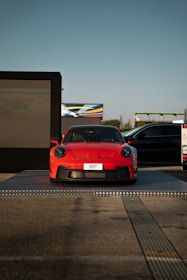 A bright red sports car is displayed on a raised platform outdoors, with a large digital screen on one side and another vehicle partially in view. The setting appears to be an automotive event or showcase, with clear blue skies in the background.