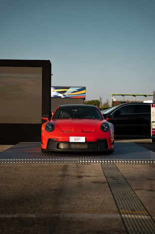 A bright red sports car is displayed on a raised platform outdoors, with a large digital screen on one side and another vehicle partially in view. The setting appears to be an automotive event or showcase, with clear blue skies in the background.
