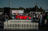 A vibrant red sports car is on display at an outdoor event, surrounded by a crowd of people. The setting includes a white picket fence and several tents or kiosks. A large, colorful backdrop with the text 'Porsche Festival 2023' is visible in the background. Trees and a clear sky frame the scene.