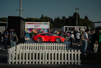 A vibrant red sports car is on display at an outdoor event, surrounded by a crowd of people. The setting includes a white picket fence and several tents or kiosks. A large, colorful backdrop with the text 'Porsche Festival 2023' is visible in the background. Trees and a clear sky frame the scene.