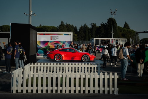 A vibrant red sports car is on display at an outdoor event, surrounded by a crowd of people. The setting includes a white picket fence and several tents or kiosks. A large, colorful backdrop with the text 'Porsche Festival 2023' is visible in the background. Trees and a clear sky frame the scene.
