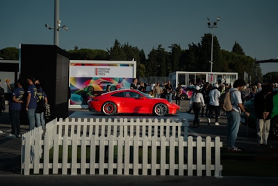 A vibrant red sports car is on display at an outdoor event, surrounded by a crowd of people. The setting includes a white picket fence and several tents or kiosks. A large, colorful backdrop with the text 'Porsche Festival 2023' is visible in the background. Trees and a clear sky frame the scene.